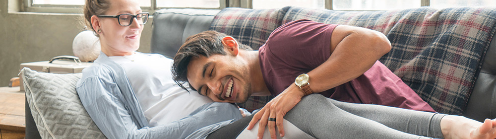 A wife laying on the couch with her husband's head on her stomach waiting for the baby to kick